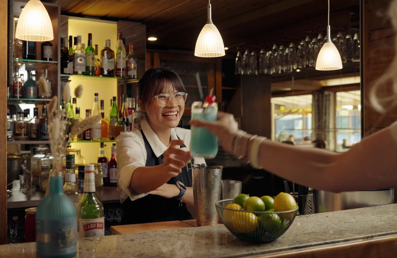 Barkeeperin reicht lächelnd einen blauen Cocktail über die Bar – Videostill, Seehotel Wiesler